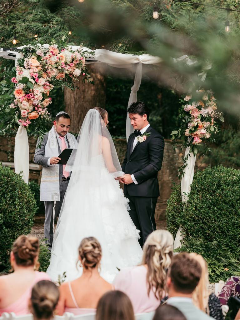 A bride and groom stand with their officiant beneath a chuppah draped in white fabric and trimmed with flowers