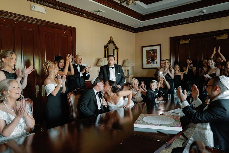 A couple kisses at their ketubah signing while family and friends applaud around the table