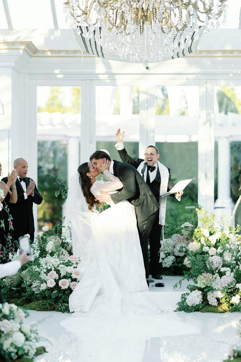 A bride and groom share their first kiss as a married couple while Todd Bronshtein cheers behind them