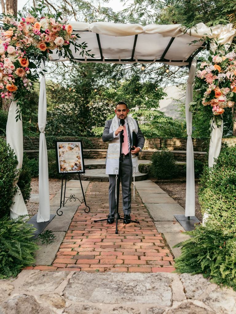 Todd Bronshtein speaking at the microphone beneath a flower-trimmed chuppah
